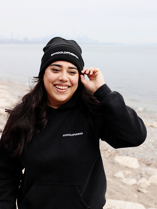 Person wearing a black knit toque and hoodie, both with the words School of Design written across the front in white embroidery. Person is adjusting toque.