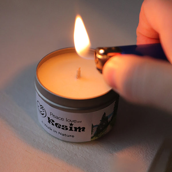 hand lighting small white candle in silver container. Lid is off and leaning on side of candle. Text on package reading "A Walk in Nature"