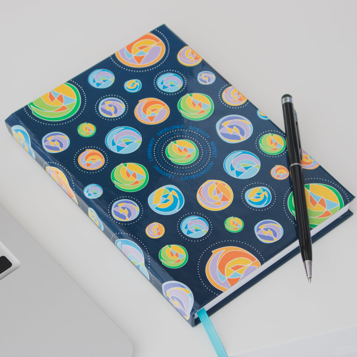 close up of 5.25" x 7.25" hardcover navy notebook with multicolour circular design pattern featuring shades of green, purple, blue, yellow and black george brown college pen