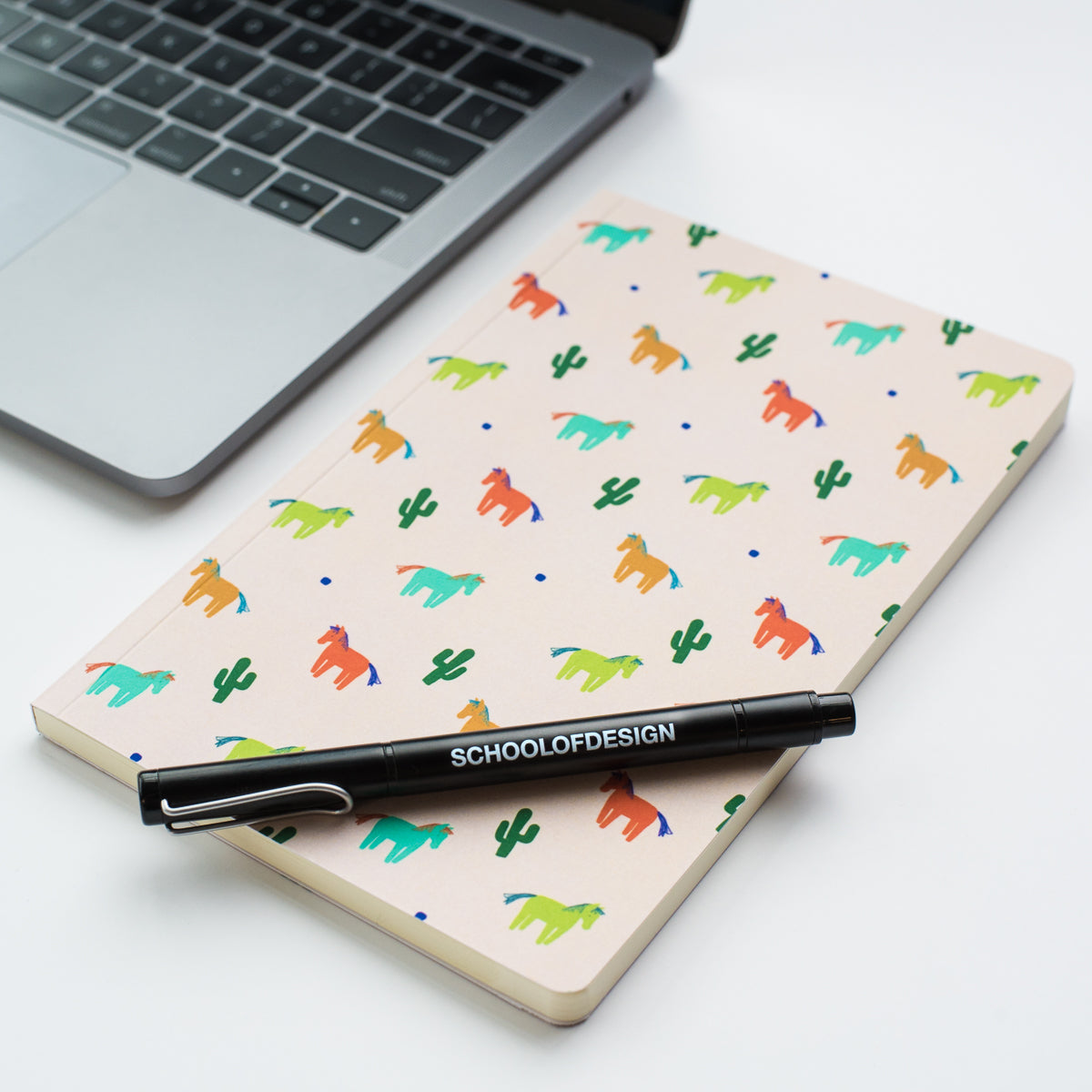 close up of 5" x 8.25" soft cover notebook with cactus and horse illustration pattern in shades of green, yellow, blue, orange and school of design ball point pen in black