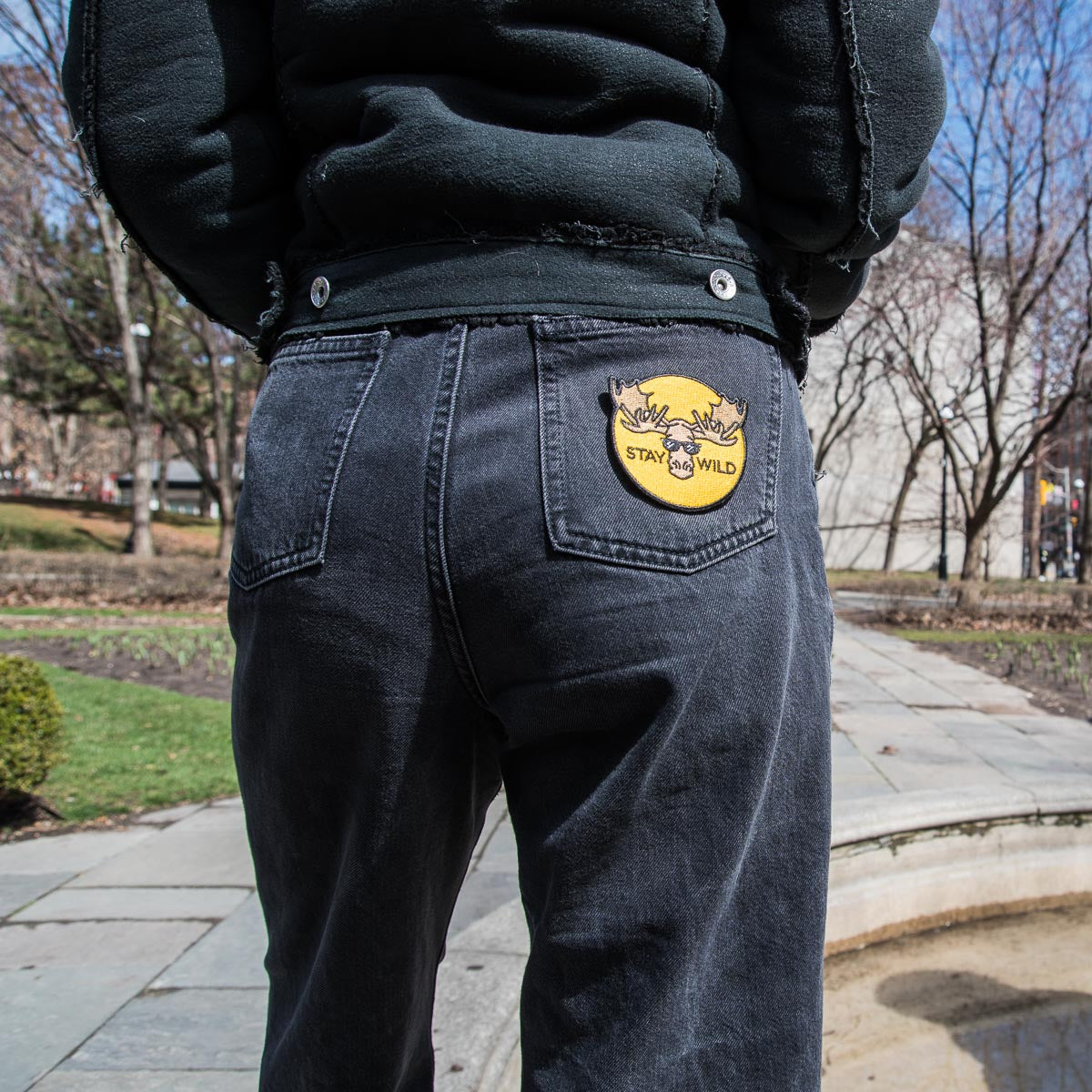 girl wearing black jeans with yellow circle patch with moose illustration with sunglasses and stay wild text featured on the right jean pocket
