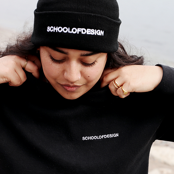 Person wearing a black knit toque and hoodie, both with the words School of Design written across the front in white embroidery. Person is adjusting collar of hoodie.
