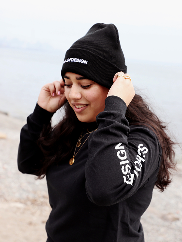 Person wearing a black knit toque with the words School of Design written across the front in white embroidery and long sleeve with the words School of Design written down the arm. Person is asjting hat.