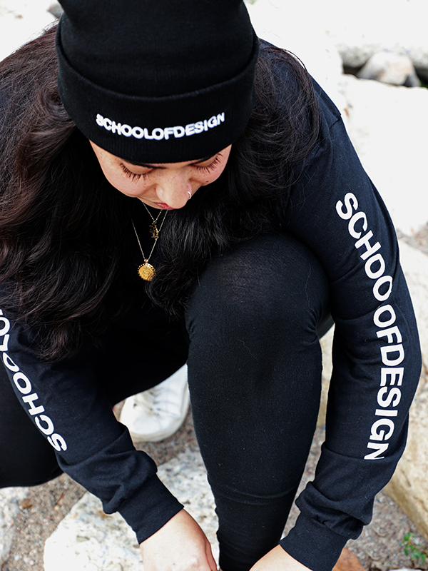 Person wearing a black knit toque with the words School of Design written across the front in white embroidery and long sleeve with the words School of Design written down the arm. Person is tying shoe.