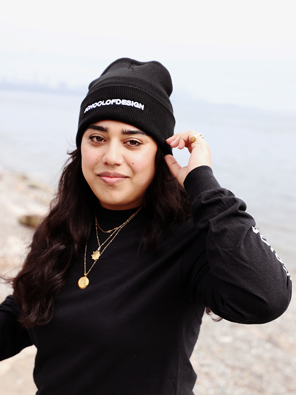 Person wearing a black knit toque with the words School of Design written across the front in white embroidery. Person is adjusting toque.