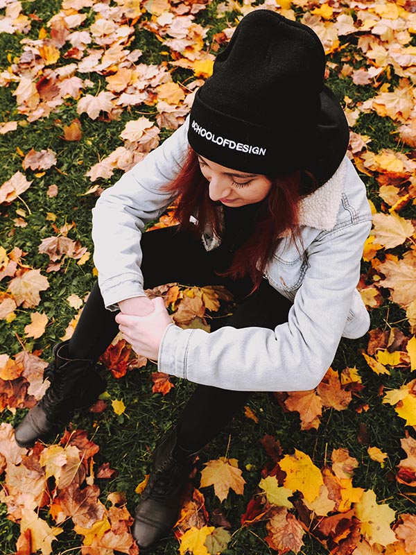 Person sitting on fall leaves on ground, wearing School of Design toque (black with white writing), jean jacket, and black hoodie