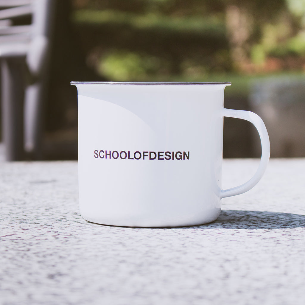 close up of white enamel mug with stainless steel rim and school of design text in black