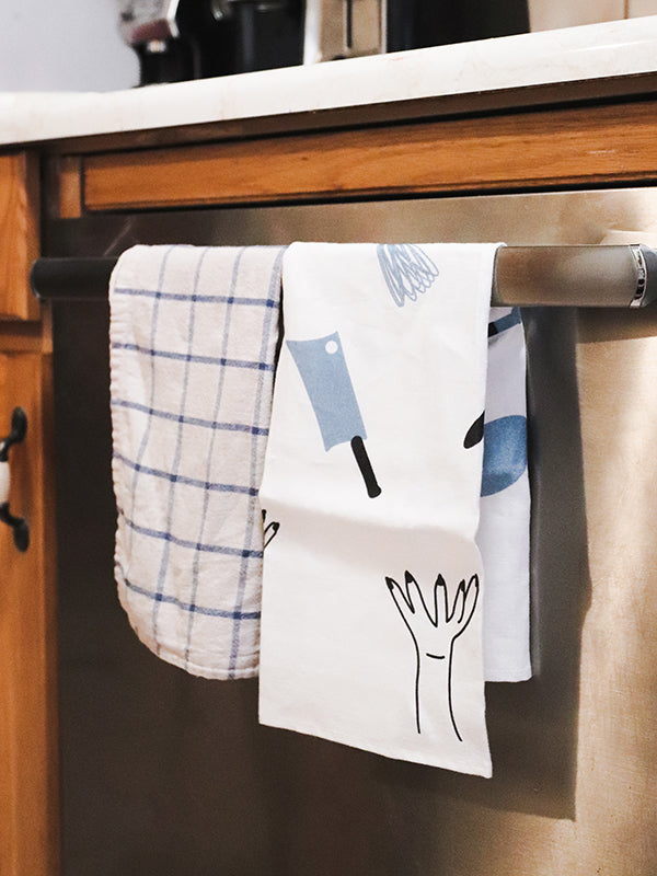 16" x 24" 100% linen tea towel featuring illustrations of various kitchen utensils and two hands tossing them into the air