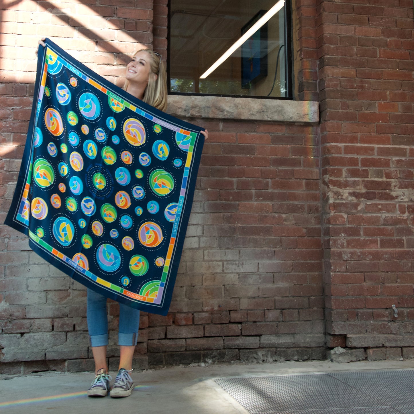 girl holding 100% silk scarf showcasing the entire pattern of circular shapes with shades of green, yellow, orange, purple and blue and matching border